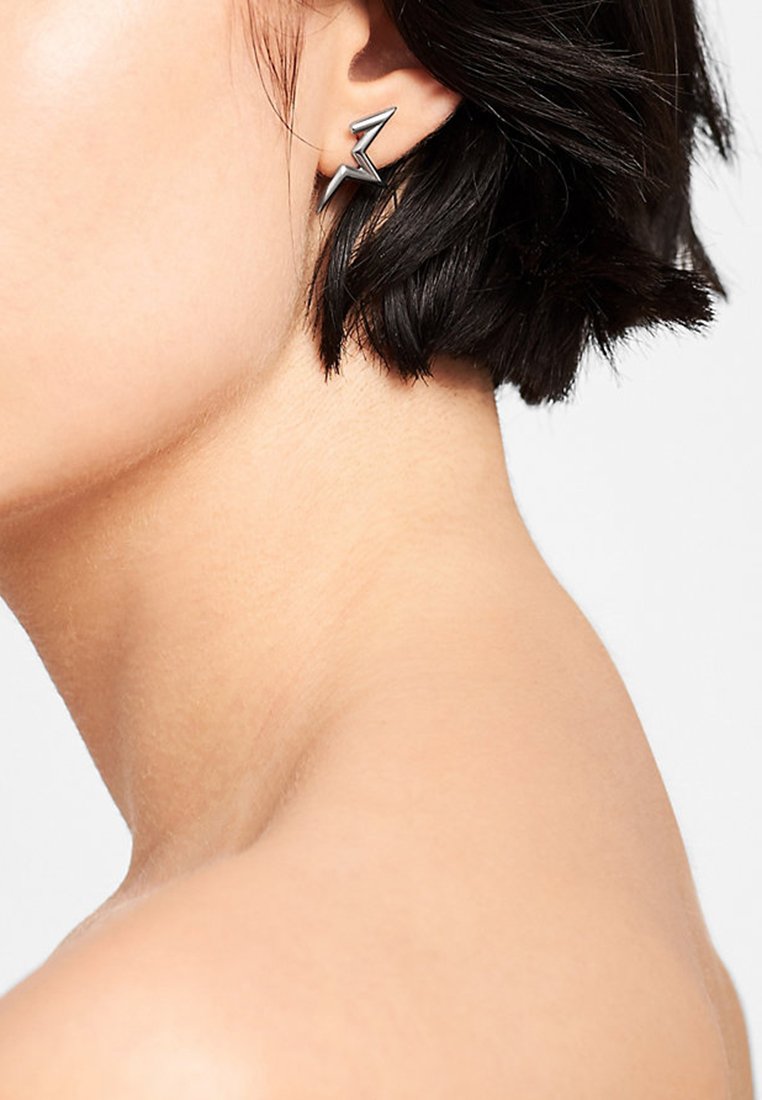 Boucles d'oreilles en métal en forme d'éclair noir. Présentent une finition polie et un design géométrique. Portées dans l'oreille.