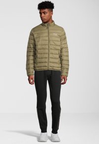 Olivgrön, vadderad jacka med hög krage och blixtlås framtill; har sidofickor, logotyp på bröstet, kombinerad med svarta byxor och vita sneakers.