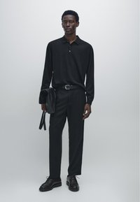 Zwarte polo met lange mouwen en een kraag met knopen, gecombineerd met een zwarte broek. Houdt een zwarte leren tas vast en draagt donkere veterboots.