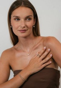 Parel oorbellen en gelaagde gouden kettingen. Huidskleur is warm; de sieraden hebben gladde, glanzende texturen en delicate ontwerpen. Lichtbruine stof.