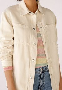 Giacca in denim beige con colletto, bottoni a scatto, due tasche sul petto e polsini a coste con dettagli pastello, indossata sopra un top in maglia a righe.