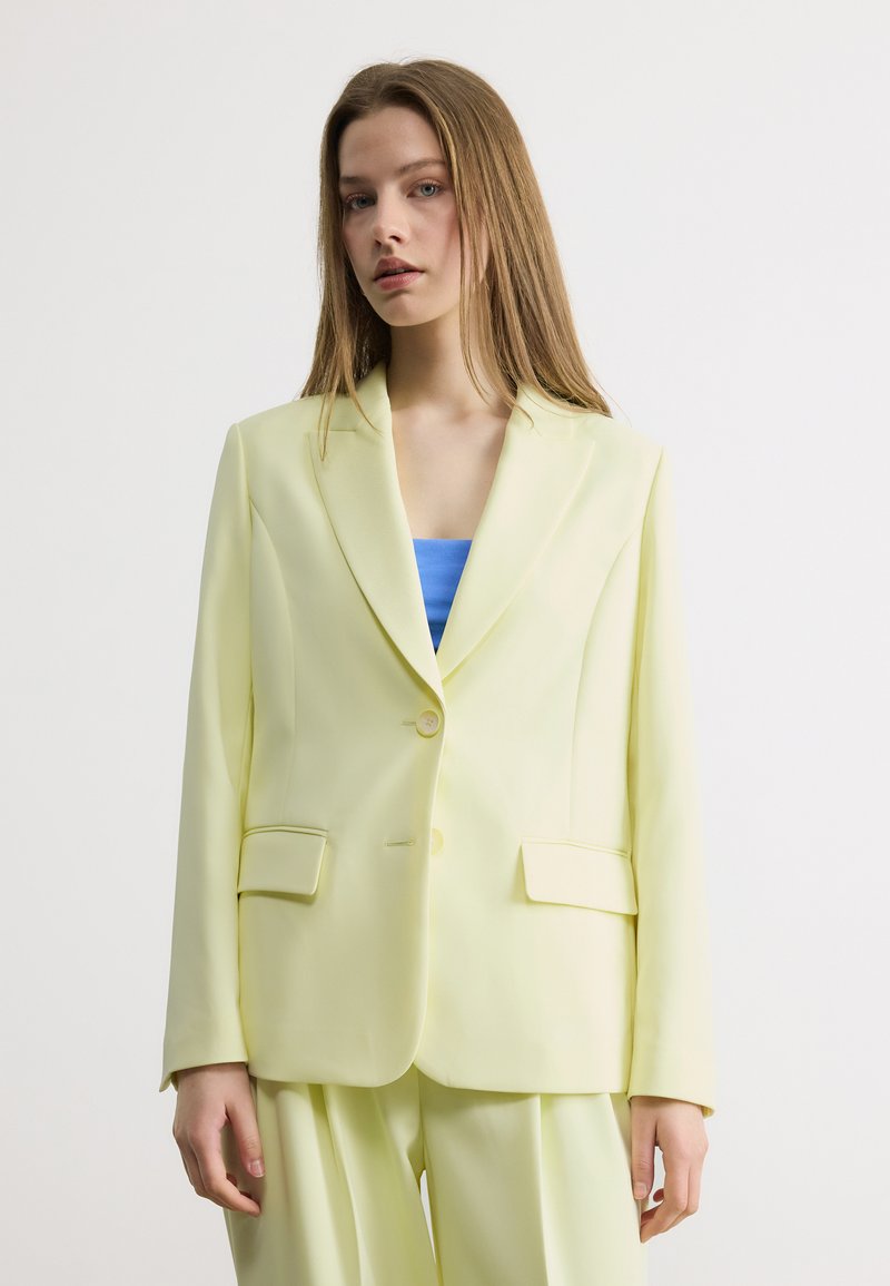 Jeune femme aux longs cheveux portant une veste de costume jaune pâle et un pantalon assorti sur un haut bleu, sur un fond uni.