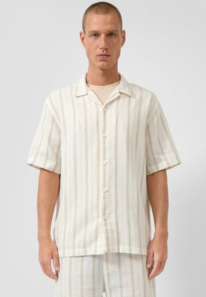 Mężczyzna ubrany w kremową, krótkorękawową koszulę na guziki z cienkimi pionowymi beżowymi paskami oraz pasujące szorty na jasnym, jednolitym tle.