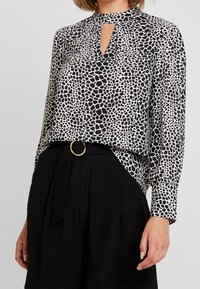 Blouse à motifs noir et blanc avec un décolleté en forme de clé, des manches longues et une coupe décontractée, accompagnée d'un pantalon noir taille haute avec une ceinture en anneau doré.