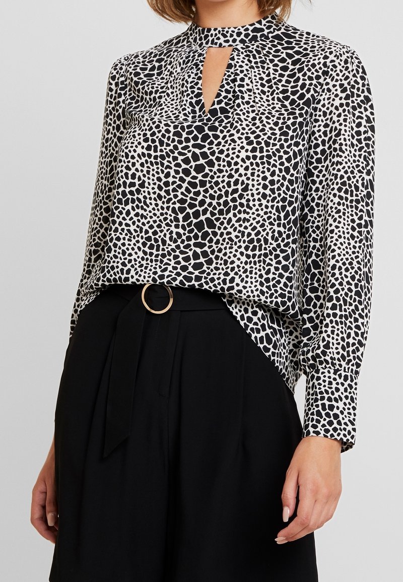 Blouse à motifs noir et blanc avec un décolleté en forme de clé, des manches longues et une coupe décontractée, accompagnée d'un pantalon noir taille haute avec une ceinture en anneau doré.