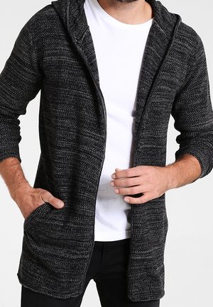 Mężczyzna w czarno-szarej, teksturowanej bluzie rozpinanej na zamek, na białej koszulce, z jedną ręką w kieszeni, a drugą regulującą zamek.