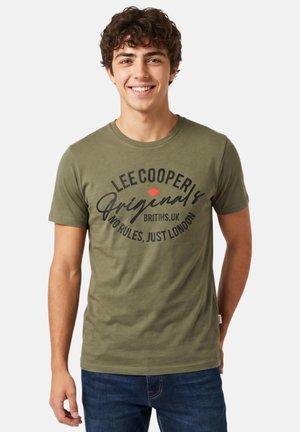 Jeune homme portant un t-shirt vert olive Lee Cooper Originals avec le texte « No rules, just London », souriant devant un fond clair uni.