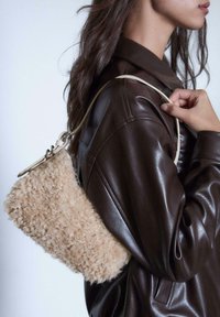 Borsa in pelliccia beige con una texture morbida, sostenuta da una tracolla chiara, indossata su una giacca di pelle marrone.