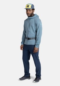 Hellblau, Reißverschlussjacke mit Kapuze, grauer Gürtel. Marineblaue Hose, graue Sportschuhe. Kappe mit blauem, gelbem und schwarzem Farbblockdesign.
