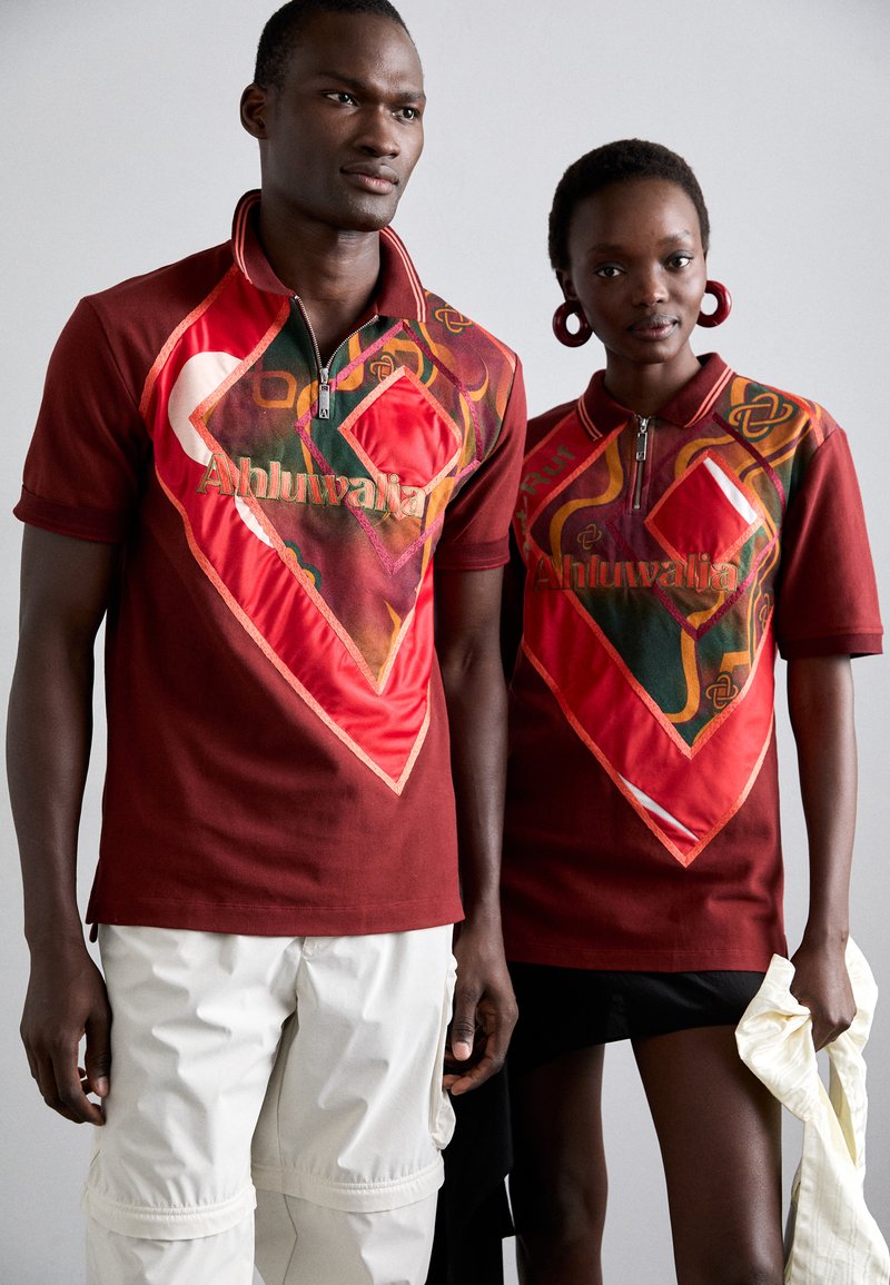 Twee personen dragen rode polo-shirts met korte mouwen, voorzien van geometrische patronen, gelaagde texturen en ritsen. De ontwerpen hebben contrasterende kleuren en logo's.