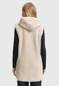 Beige Fleece-Weste mit Kapuze, die eine strukturierte, weiche Oberfläche aufweist. Die Rückseite hat ein einfaches Design und reicht bis zur Mitte des Oberschenkels für zusätzliche Bedeckung.