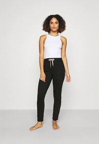 Zwarte joggingbroek met een tailleband met trekkoord, gecombineerd met een wit mouwloos topje. Het materiaal lijkt zacht en rekbaar, ontworpen voor comfort.