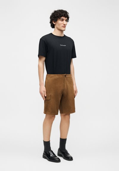 Jeune homme aux cheveux bouclés portant un t-shirt noir avec logo, un short cargo marron arrivant aux genoux, des chaussettes foncées et des chaussures en cuir noires, debout les mains le long du corps.