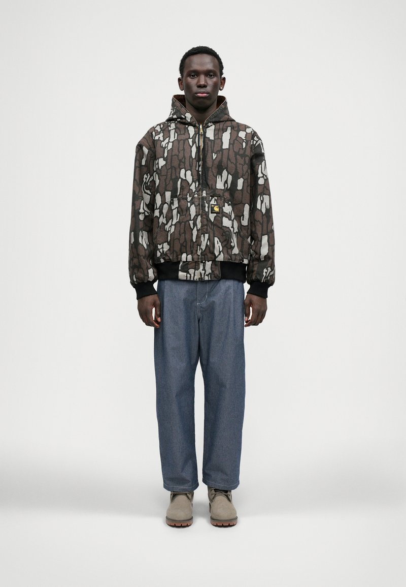 Braune und weiße gemusterte Kapuzenjacke mit vorderem Reißverschluss und Kängurutasche, kombiniert mit locker geschnittenen blauen Jeans und beigen Schuhen.