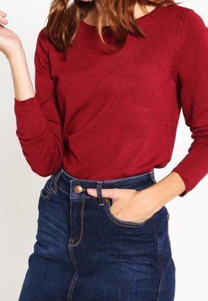 Personne portant un haut rouge ajusté à manches longues, rentré dans une jupe en jean bleu foncé taille haute avec bouton et poche sur le devant.