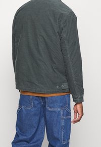 Donkergroene corduroy jas met een ronde achterkant, voorzien van knoopaccenten en een geribde bruine trui eronder, gecombineerd met blauwe spijkerbroeken.