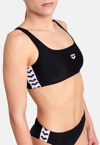 Svart bikiniöverdel med fyrkantig halsringning, med sidodetaljer i vita geometriska mönster. Slät tyg med en figursydd design.