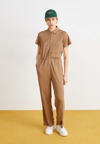 Combinaison marron côtelée avec manches courtes, fermeture à boutons sur le devant et taille élastique. Associée à des baskets blanches à plateforme et une casquette verte.