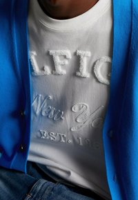 Modrý cardigan přes bílou mikinu s texturovaným nápisem "ALFIE" a "New York EST. 1985." Viditelné džíny.