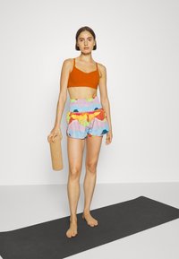 Sutiã desportivo laranja, calções de padrão multicolorido com uma cintura franzida, tapete de yoga de cortiça natural na mão, de pé sobre um tapete de yoga preto.