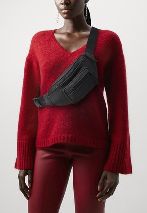 Sac banane noir décontracté avec des fermetures éclair élégantes ; porté en bandoulière sur un pull rouge texturé aux larges manches, assorti à un pantalon rouge brillant.