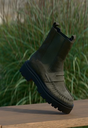 Bottine en daim vert foncé avec semelle épaisse noire à crampons reposant sur une surface en bois en extérieur avec un fond d'herbe haute et verte.