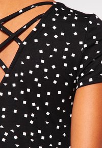 Haut noir avec des motifs carrés et triangulaires blancs. Présente un design de bretelles croisées au dos et des manches courtes. Texture de tissu douce.