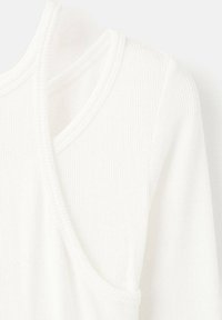 Top bianco a coste con scollatura a V e unici tagli asimmetrici sulle spalle. Tessuto morbido, leggero e con dettagli di cucitura discreti.