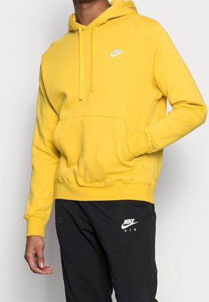 Mężczyzna w jaskrawym żółtym kapturze Nike z przednią kieszenią oraz czarnych spodniach dresowych Nike Air na białym tle.