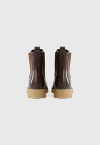 Botas de tobillo de cuero marrón con paneles elásticos laterales, tiradores, suelas gruesas beige y textura suave, mostradas desde atrás sobre un fondo blanco.