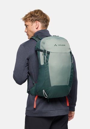 Jonge man met een groene en lichtgrijze wandelrugzak met gevoerde banden en rode ritstrekken, zijwaarts kijkend in buitenkleding.