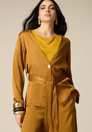 Donna che indossa una giacca giallo senape con cintura sopra un top abbinato, orecchini dorati a forma di stella e braccialetti spessi dorati e neri al polso.