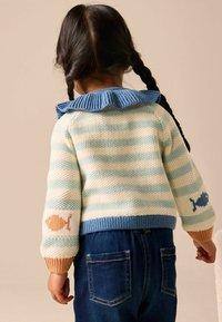 Gebreide trui met lichtblauwe en crèmekleurige strepen, een blauwe gerimpelde kraag en vispatronen op de mouwen. Gecombineerd met donkerblauwe denim.