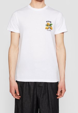 T-shirt blanc avec un petit motif de citron et le texte "IT'S OK to be different" sur le côté gauche de la poitrine, porté avec un pantalon noir ample.