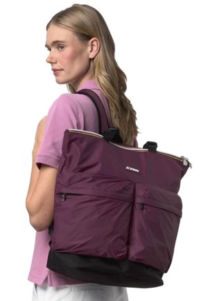 Donna con capelli biondi lunghi che indossa una camicia viola e porta una grande borsa a tracolla bordeaux e nera con tasche frontali con zip sulla spalla.