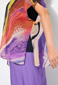Blusa trasparente multicolore con motivi astratti in arancione, viola e nero; presenta maniche ampie e fessure laterali. Abbinata a una gonna viola.