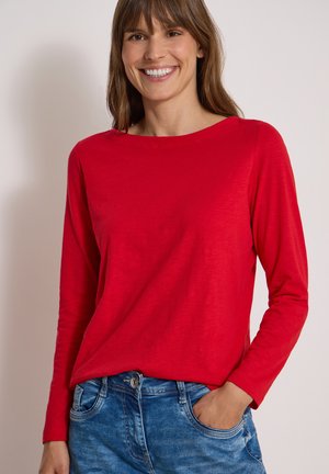 Femme souriante portant une chemise rouge vif à manches longues et un jean bleu, une main dans la poche, sur un fond uni.