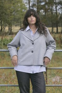 Giacca grigia in misto lana con colletto a revers, bottoni frontali e vestibilità ampia, indossata sopra una camicia a righe blu e bianche e pantaloni scuri.