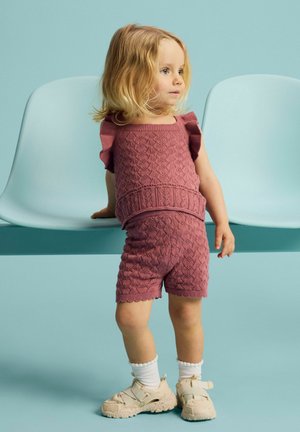 Lille barn med blondt hår iført en matchende tekstureret lyserød strikket top og shorts, hvide sokker og beige sneakers, stående ved lyseblå stole.