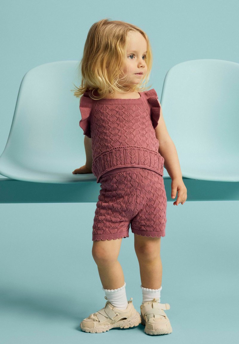 Jeune enfant aux cheveux blonds portant un haut tricoté rose texturé assorti et un short, des chaussettes blanches et des baskets beiges, debout près de chaises bleu clair.