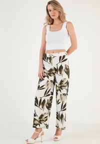 Wijde broek met een loofmotief in groen, goud en blauw op een witte achtergrond, gecombineerd met een aansluitende witte crop top en sleehakken.