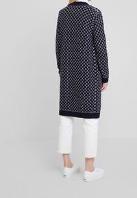 Knälång marinblå kardigan med ett vitt hjärtat mönster, ribbade ärmslut och fåll, bärs över vita cropped byxor och vita sneakers.