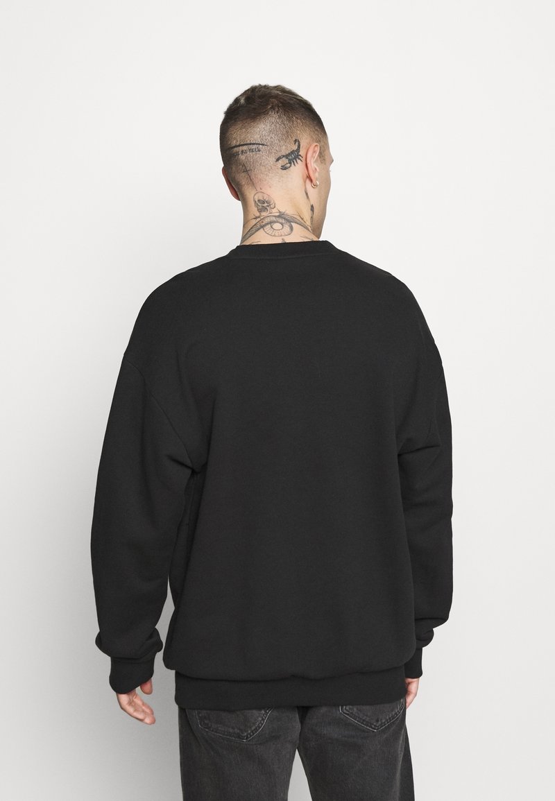 Man med kort hår och tatueringar på nacke och huvud, iförd en svart sweatshirt och mörka jeans, vänd bort från kameran mot en vit bakgrund.
