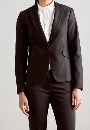 Person iført en tætsiddende sort blazer med en enkelt knap over en hvid skjortekrave og matchende sorte bukser, stående mod en ensfarvet baggrund.