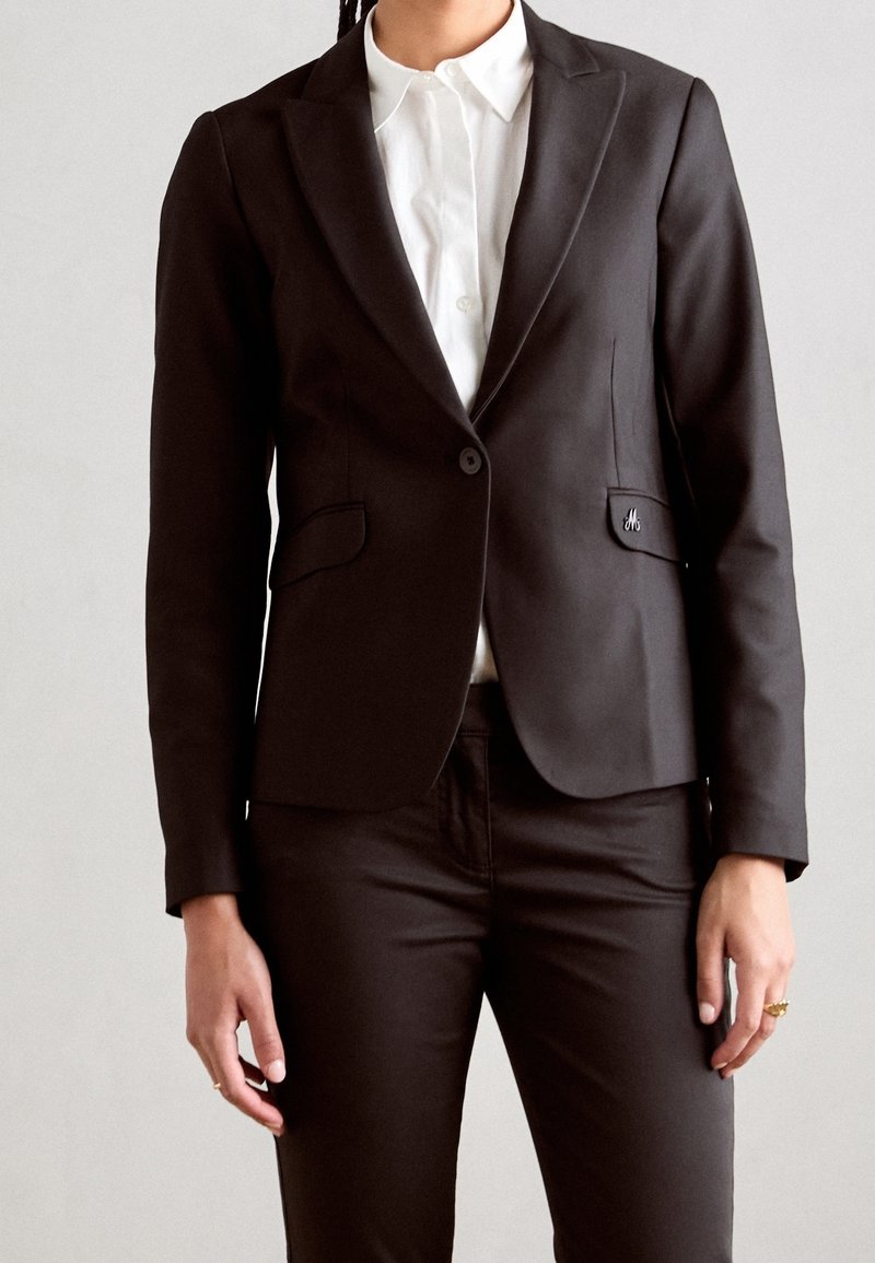 Personne portant un blazer noir ajusté avec un seul bouton sur une chemise blanche à col et un pantalon noir assorti, debout devant un fond uni.