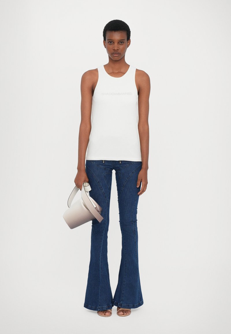 Top blanco sin mangas con la palabra "SHADOWBANNED", jeans acampanados azules y un bolso suave de color crema con un asa curva.