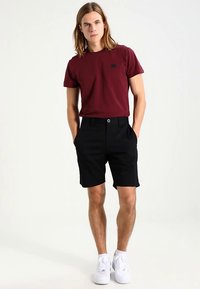 Bordeauxrode T-shirt met een klein zwart logo, gecombineerd met zwarte korte broek en witte sneakers. De outfit heeft een eenvoudig, casual ontwerp.