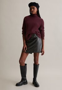 Burgunderfarbener Waffelstrickpullover, schwarzer Lederwickelrock und kniehohe schwarze Stiefel. Das Outfit hat eine strukturierte Oberfläche und eine figurbetonte Silhouette.