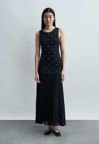 Žena stojaca, oblečená v čiernom tielku bez rukávov s malými bielymi krížikmi a čiernej plisovanej maxi sukni s čiernymi balerínkami.