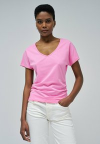 Camiseta rosa de algodón suave con cuello en V, mangas cortas y detalle sutil en el borde, combinada con pantalones blancos. Textura suave y ajuste casual.